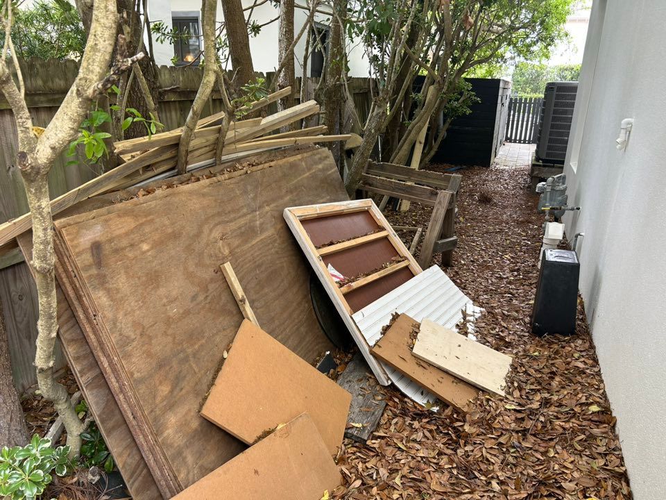 Pile of discarded wood, boards, and a window frame next to a narrow walkway lined with trees and a building.