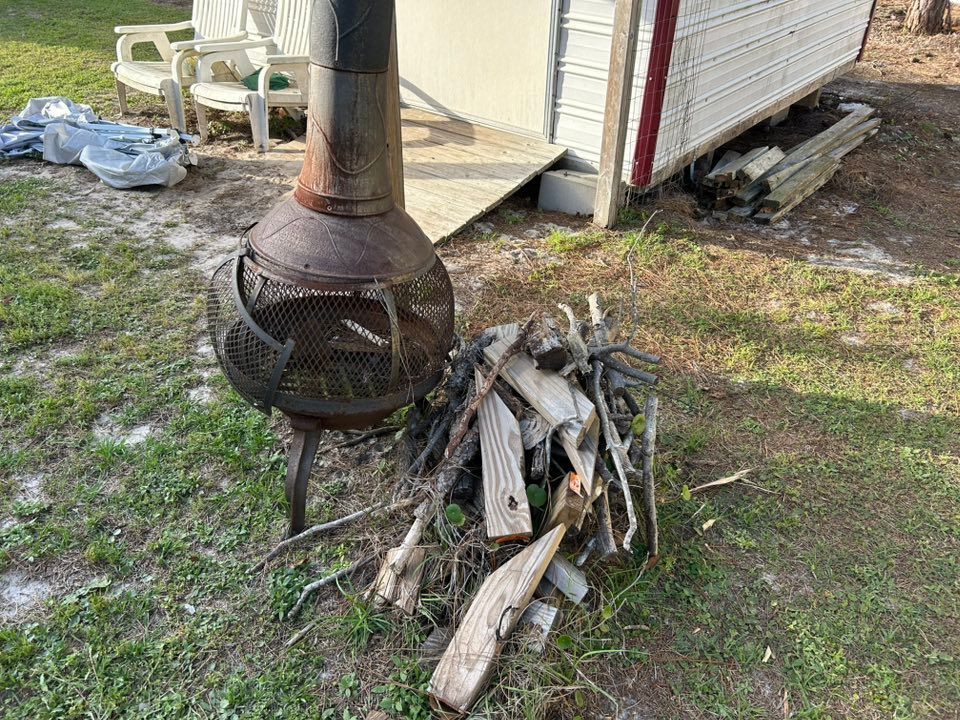 Rusty metal fire pit with pile of firewood next to a white trailer on a grassy area.