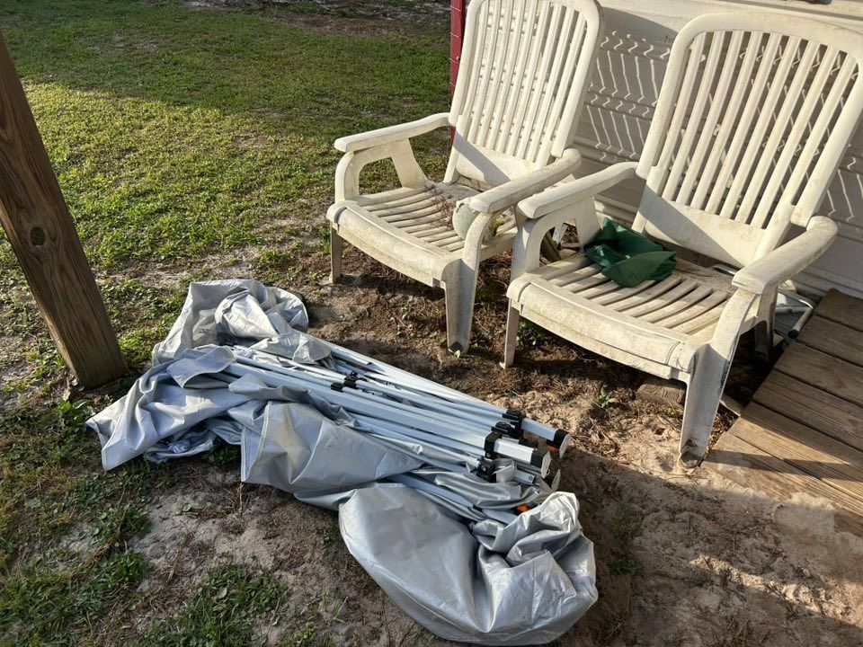 Two white plastic chairs near a tarp and folded metal frame on dirt and grass.