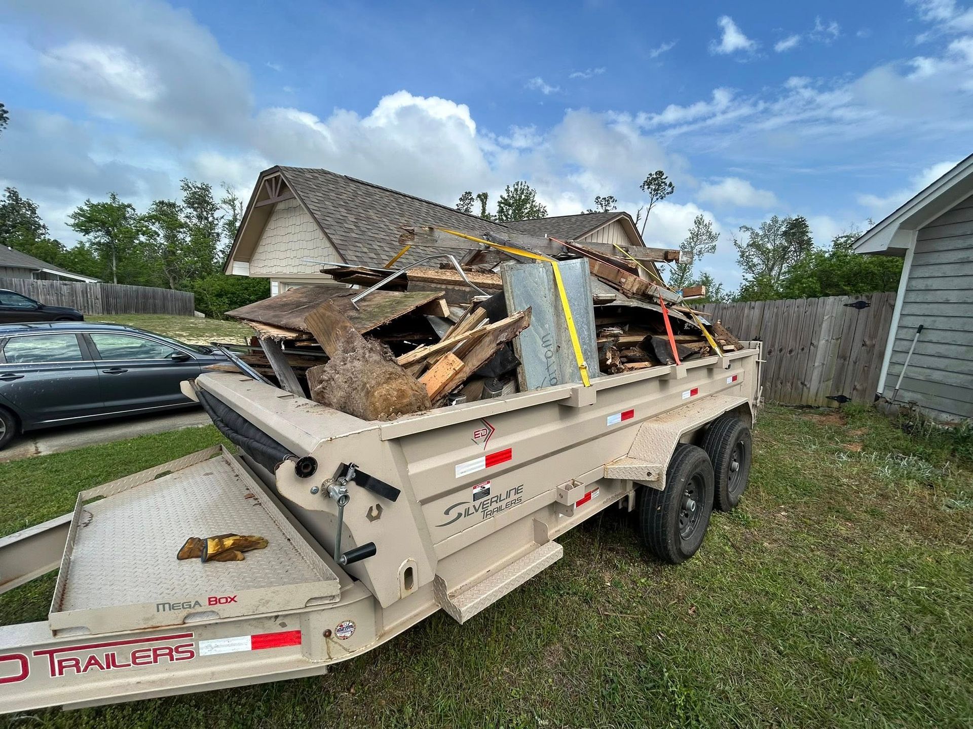Tan trailer loaded with construction debris on green grass. House and fence in background.