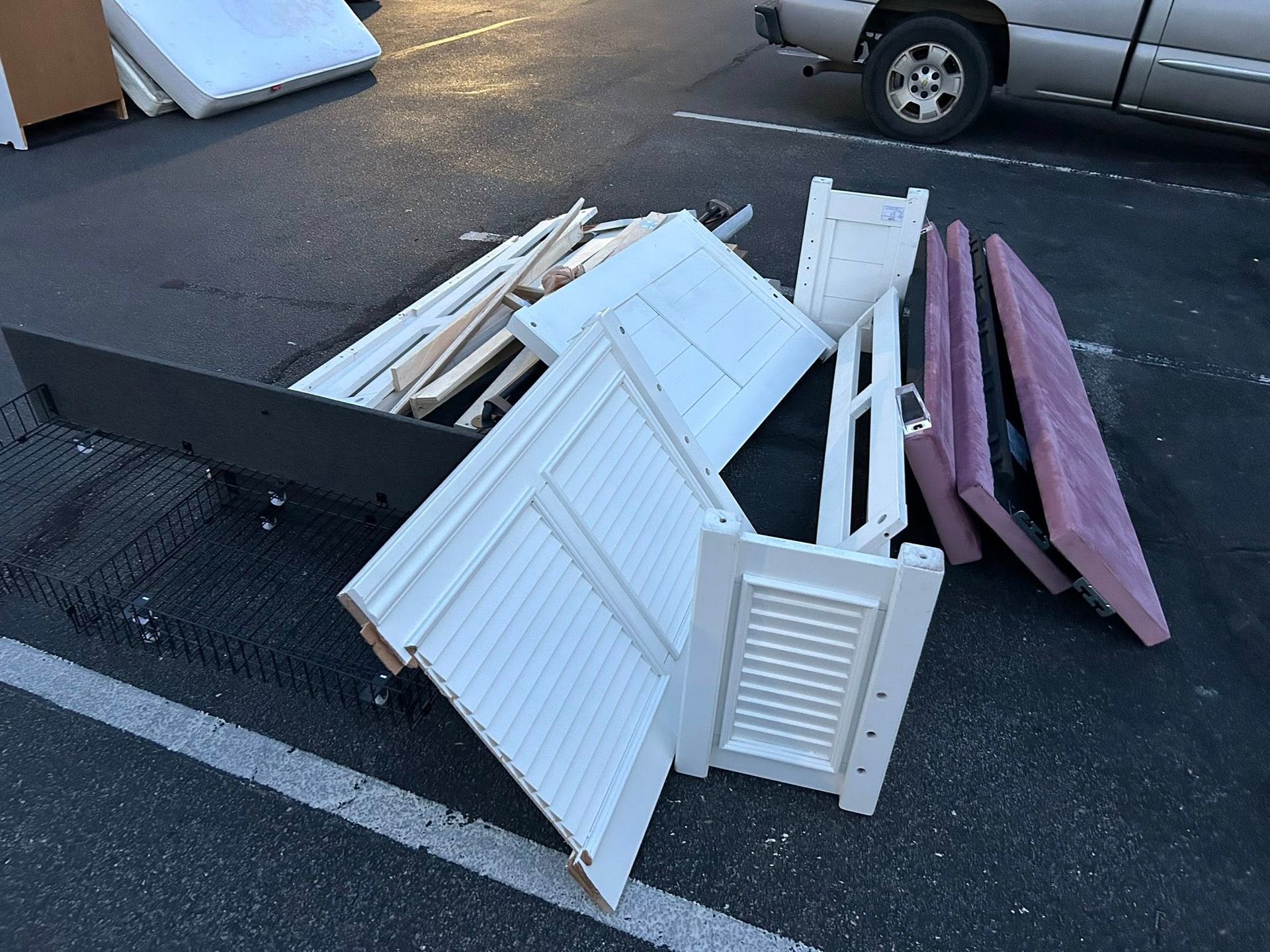 Pile of discarded white shutters, wood pieces, and pink insulation on asphalt. Silver truck in background.