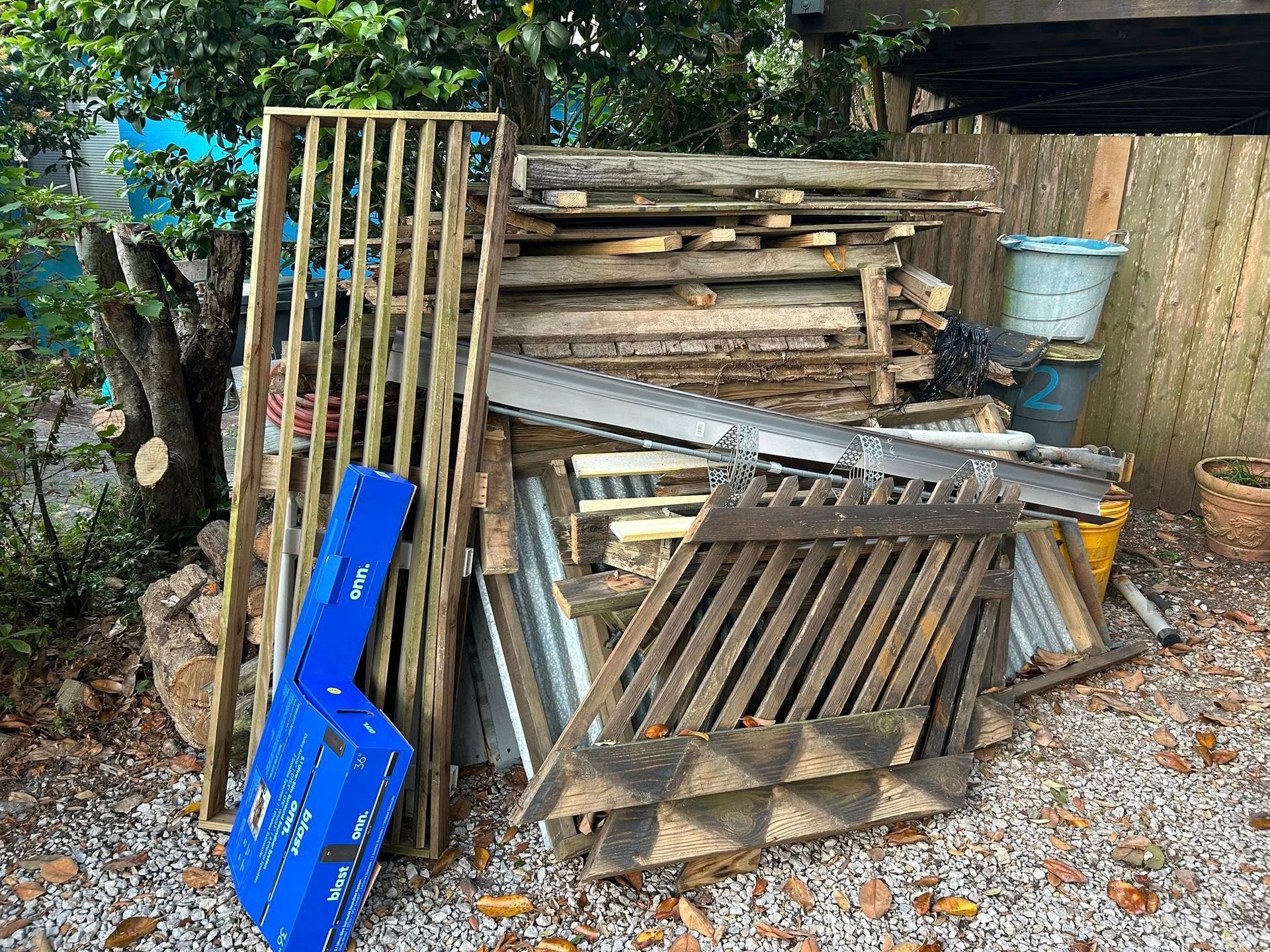 Pile of discarded lumber and metal scraps in a gravel yard, with a blue container and a wooden fence.