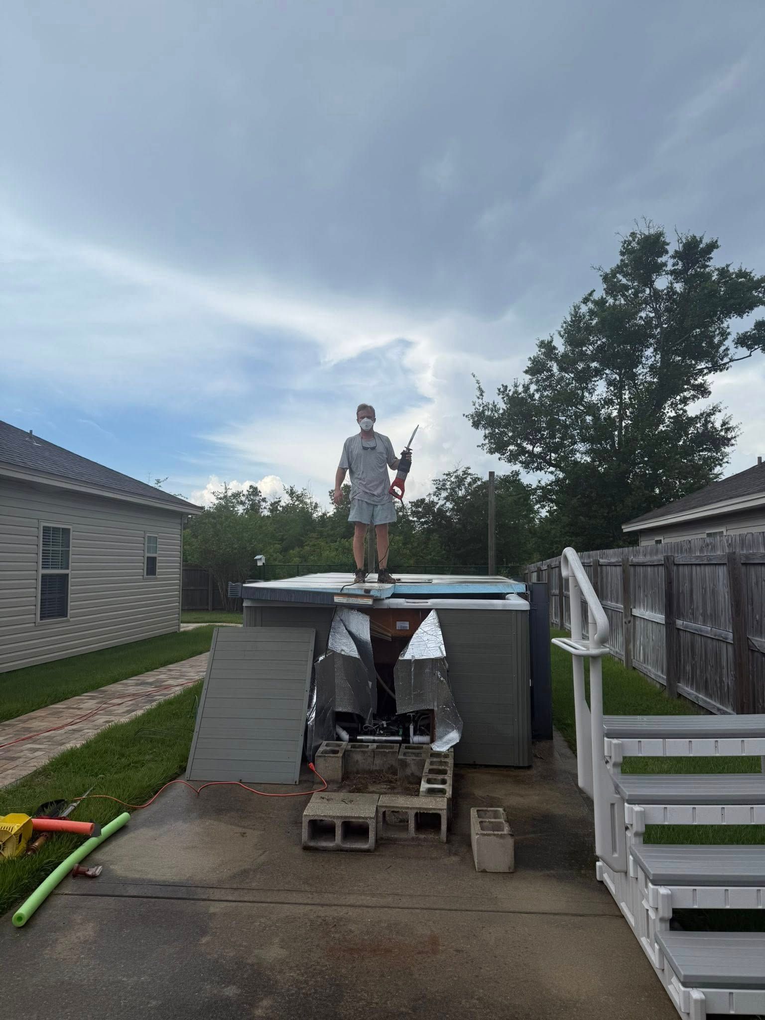 Person stands on a hot tub roof holding an object. Outdoor setting with cloudy sky and houses.