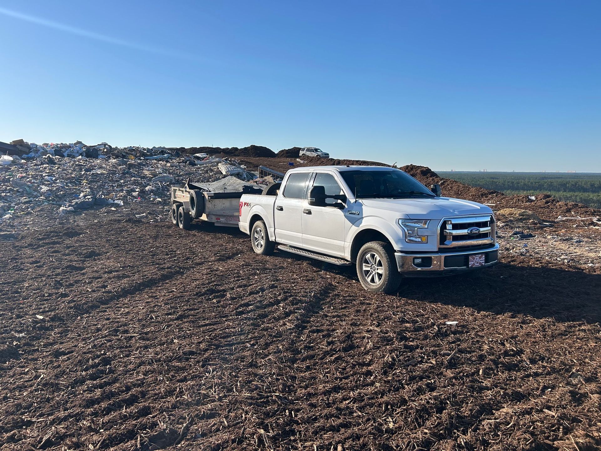 White pickup truck towing a trailer on a brown, rocky ground.  A large pile of debris is behind them under a blue sky.