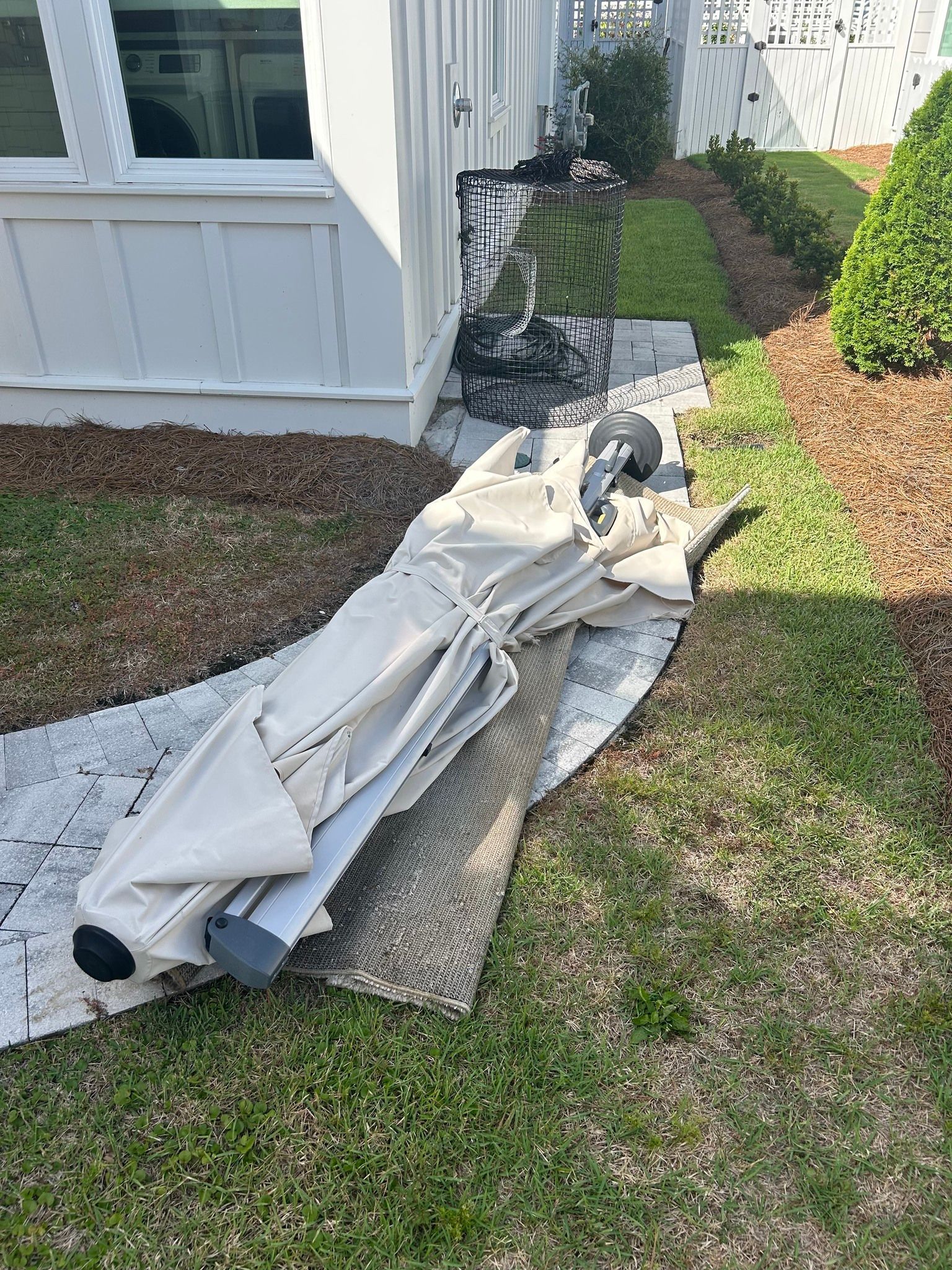A collapsed beige patio umbrella on a brick and grass pathway near a white building.