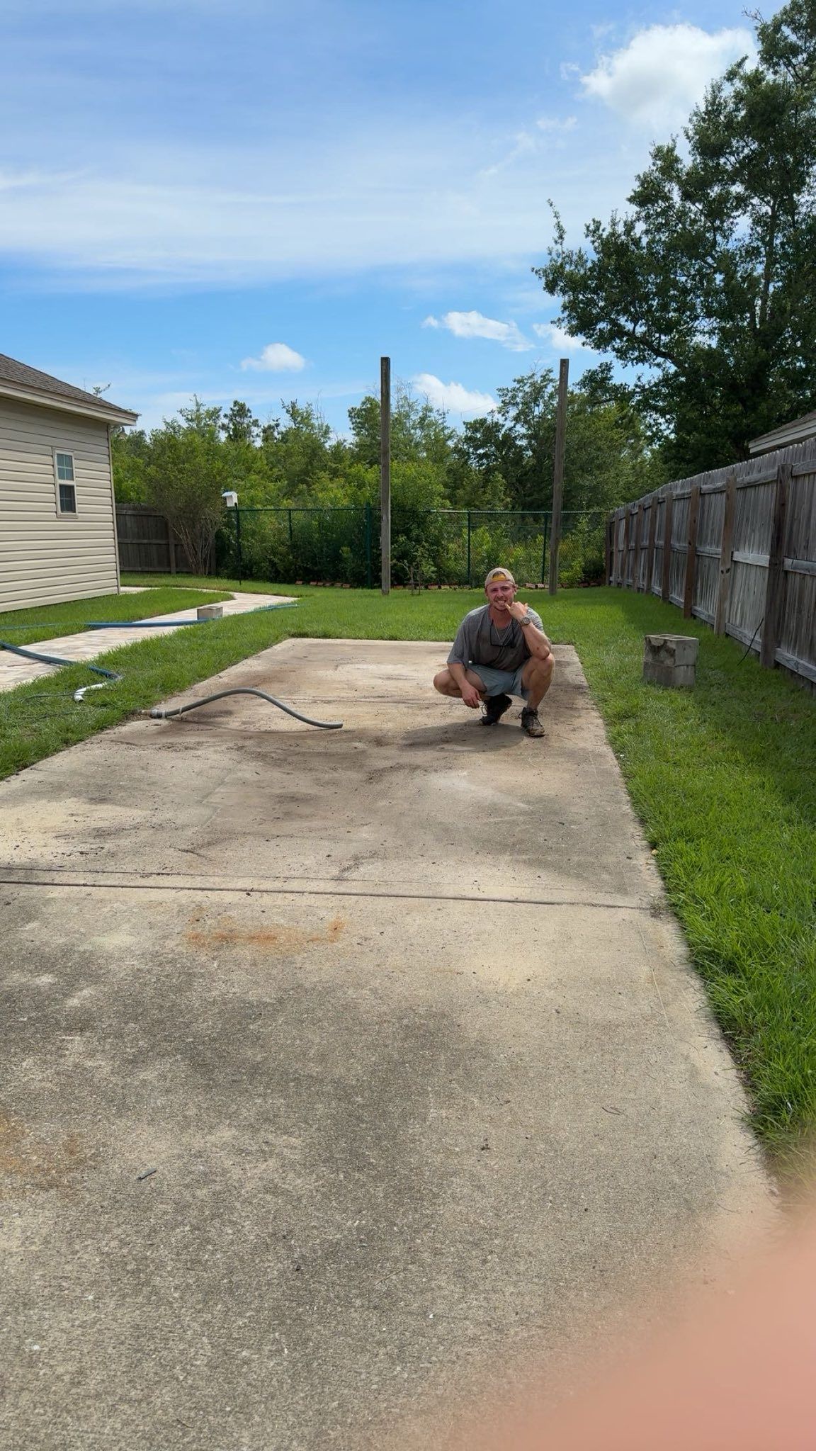 Man squatting on a concrete slab outdoors. Green grass, wooden fence, and blue sky in background.