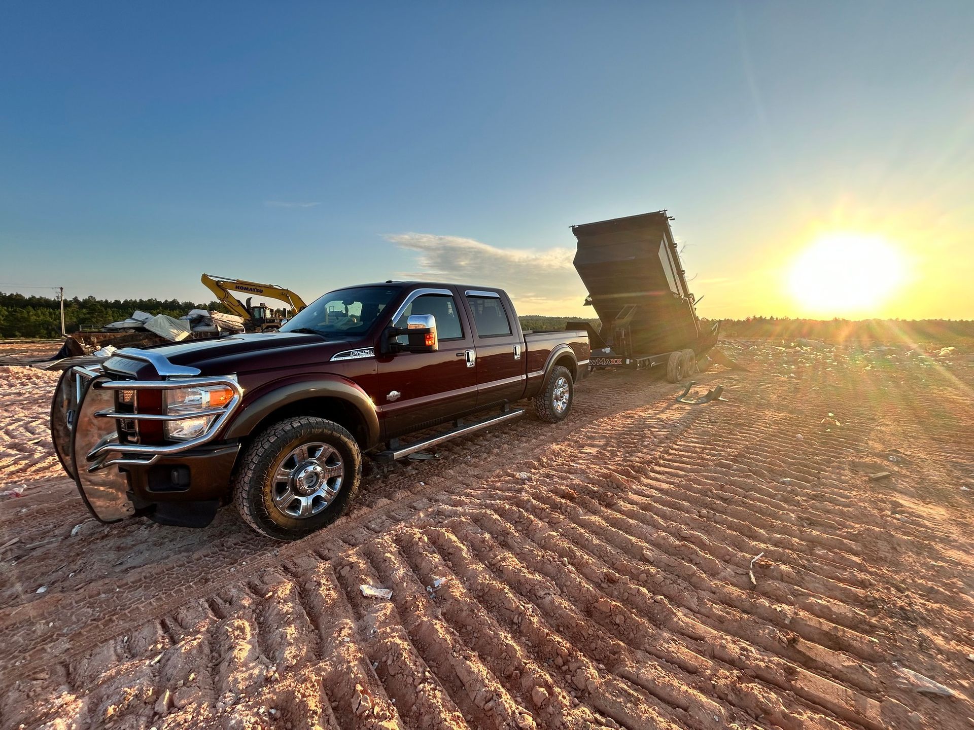 Dark red pickup truck towing a raised dump trailer on a dirt road at sunset.