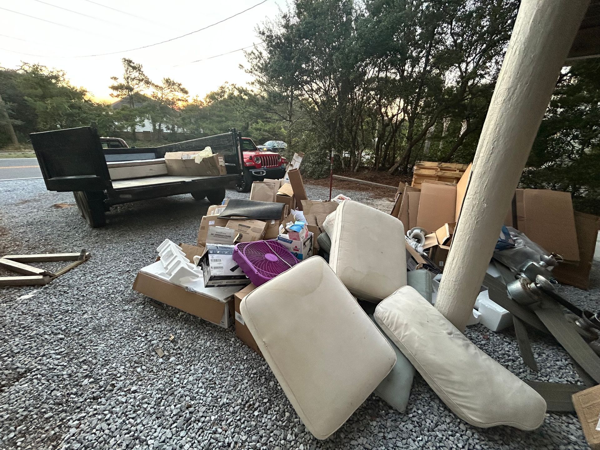 Pile of discarded household items, including cushions and cardboard boxes, next to a trailer. Outdoor setting.