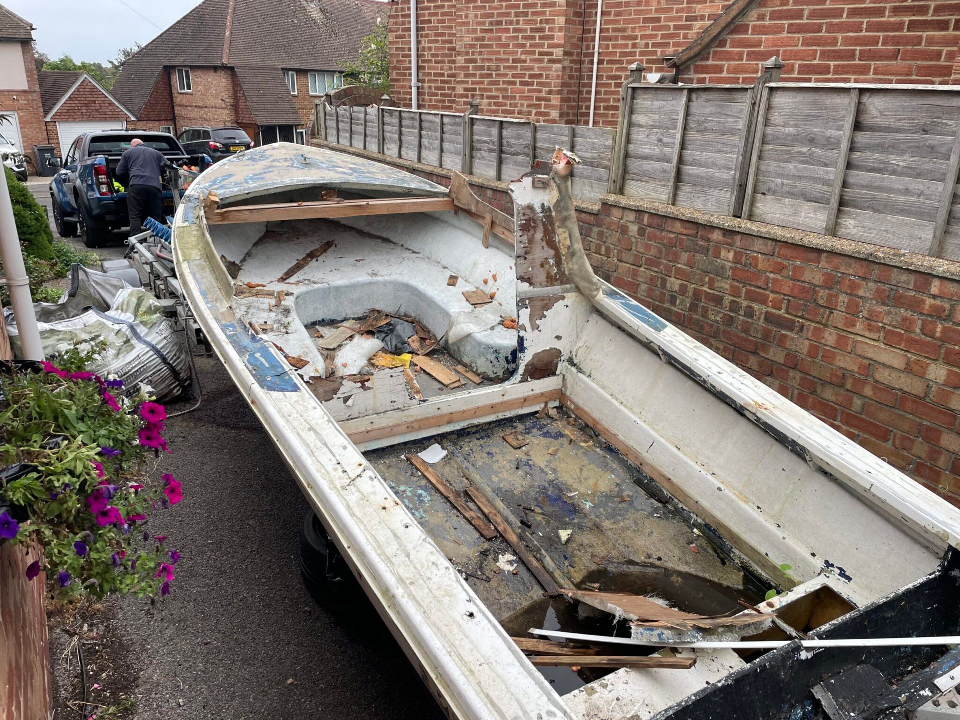 A weathered, gutted white boat in a driveway beside a brick wall, with debris scattered around.