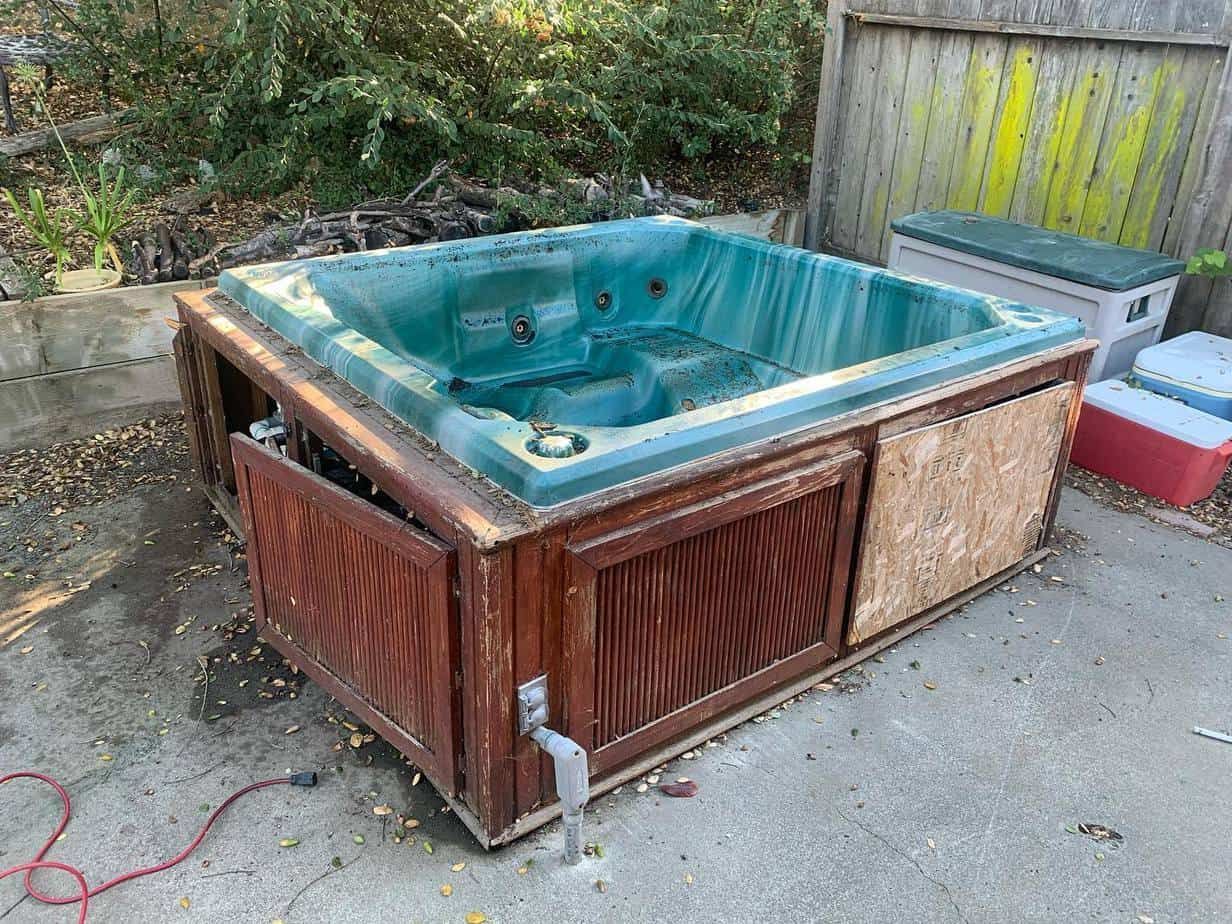 A weathered, green hot tub with a damaged wooden exterior, outdoors on concrete.