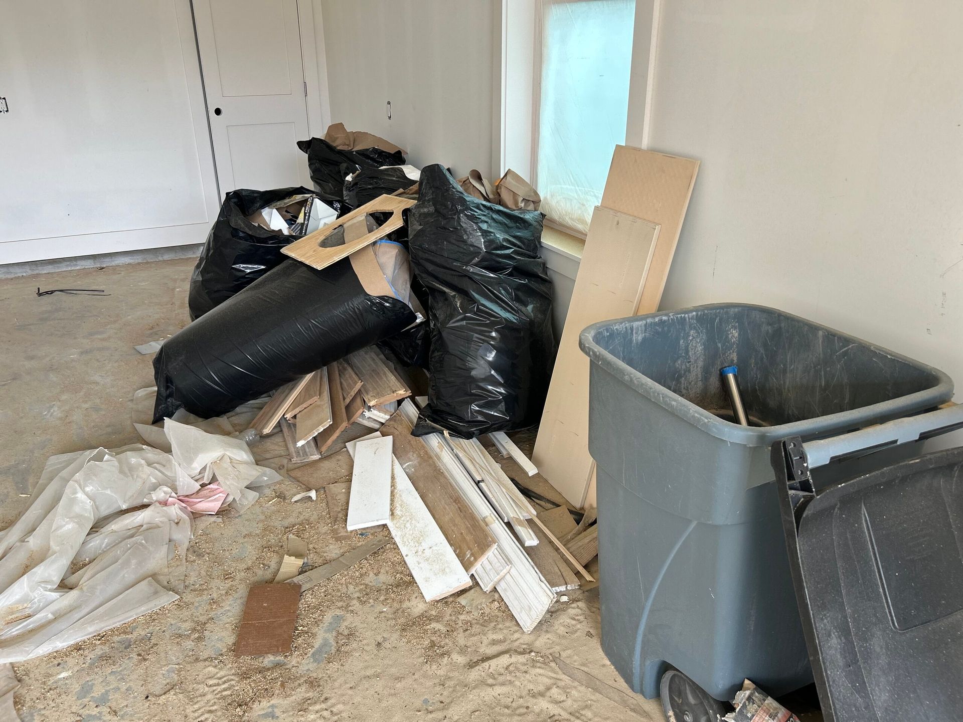 Room with construction debris: trash bags, wood, and a gray bin near a window.