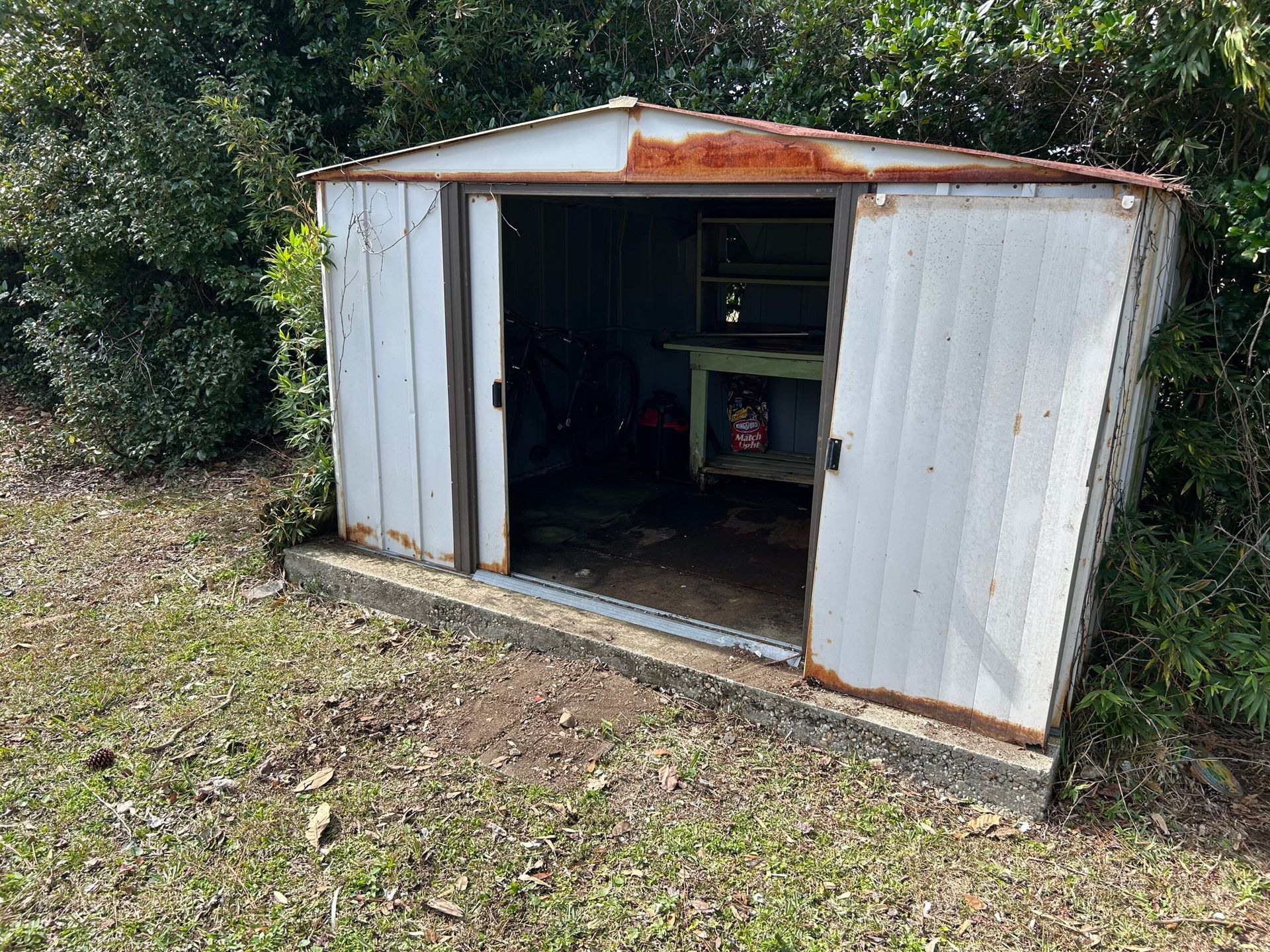 Weathered metal storage shed with open sliding doors, surrounded by grass and foliage.