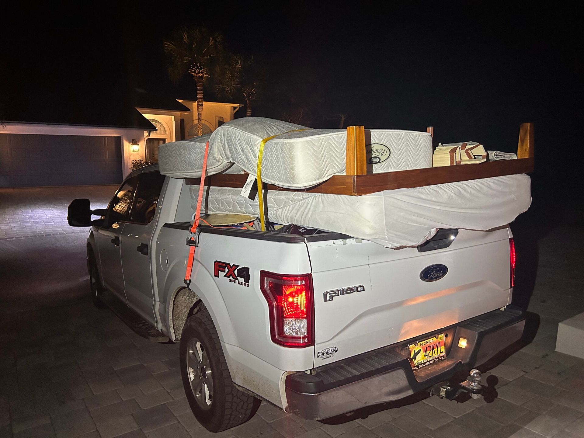 White pickup truck loaded with mattress and bed frame, secured with straps, at night.