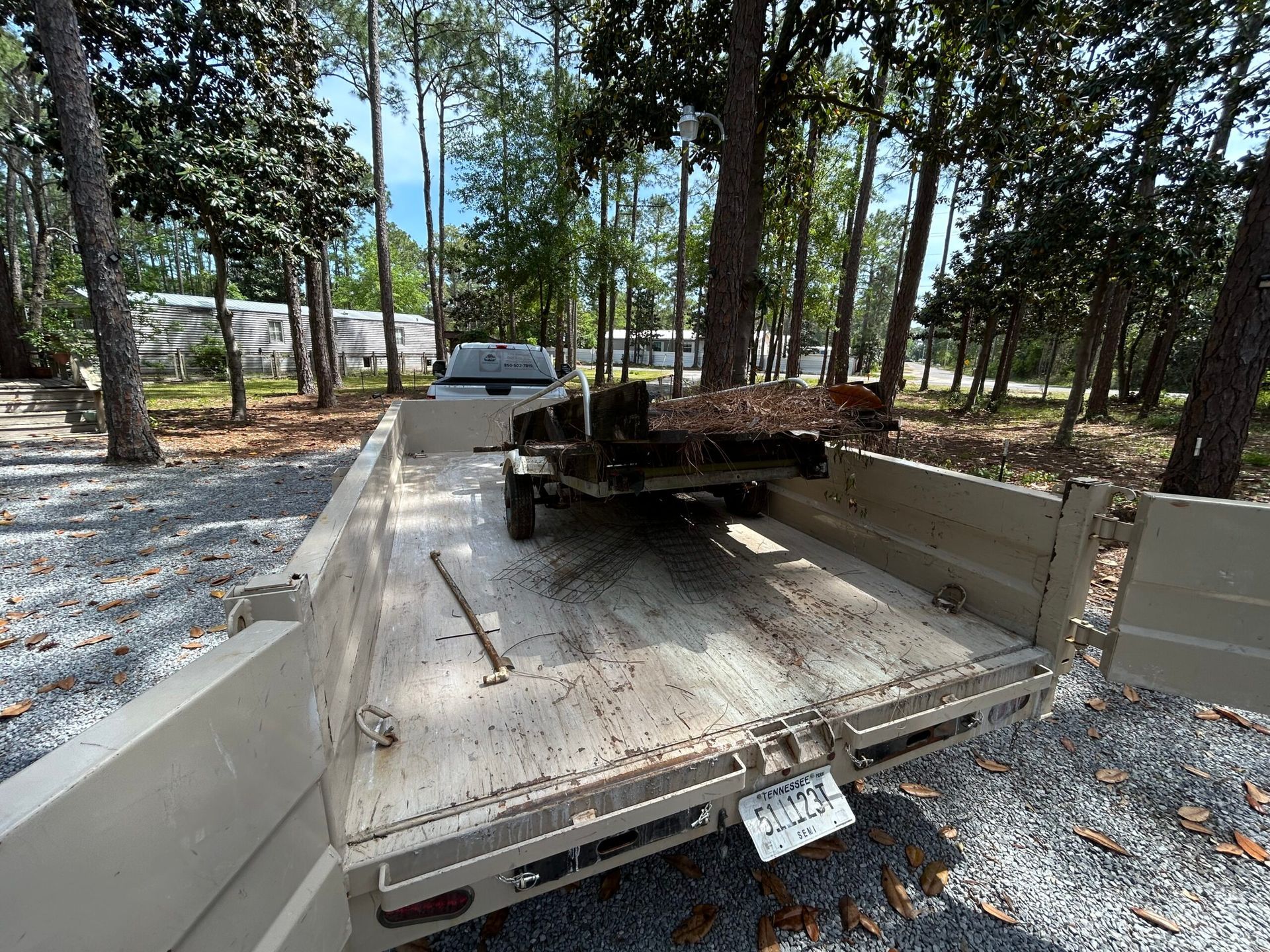 Tan flatbed trailer with a small trailer loaded, parked on gravel, surrounded by trees.