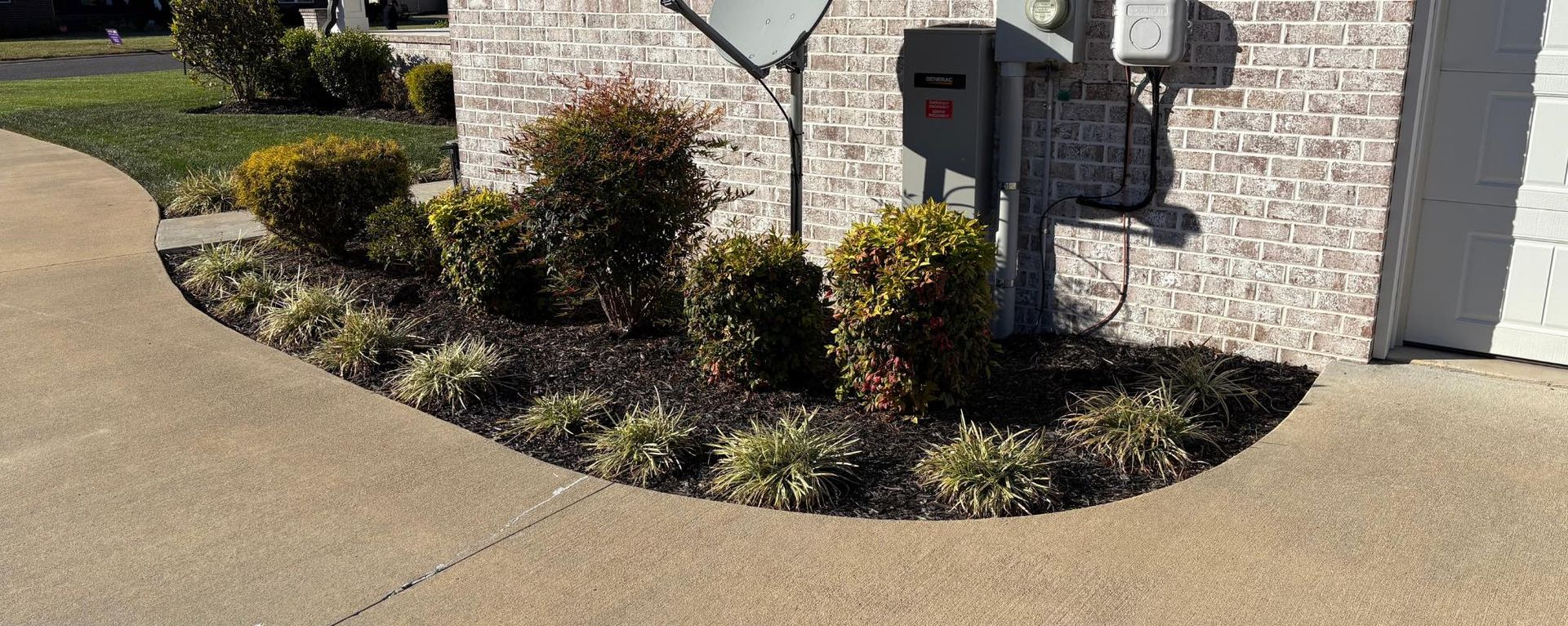 A landscaped bed with shrubs and small plants, bordering a sidewalk, in front of a brick building.