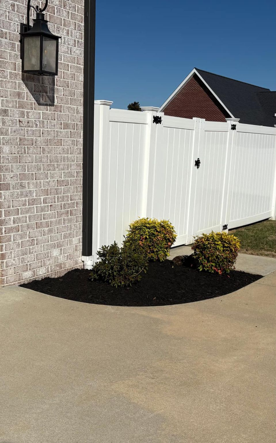 White fence with dark mulch bed, small bushes, brick wall, and blue sky.