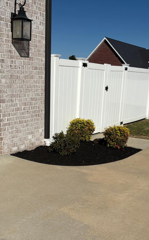 White vinyl fence with a gate, next to a brick wall with a lantern, and landscaping with mulch and bushes.