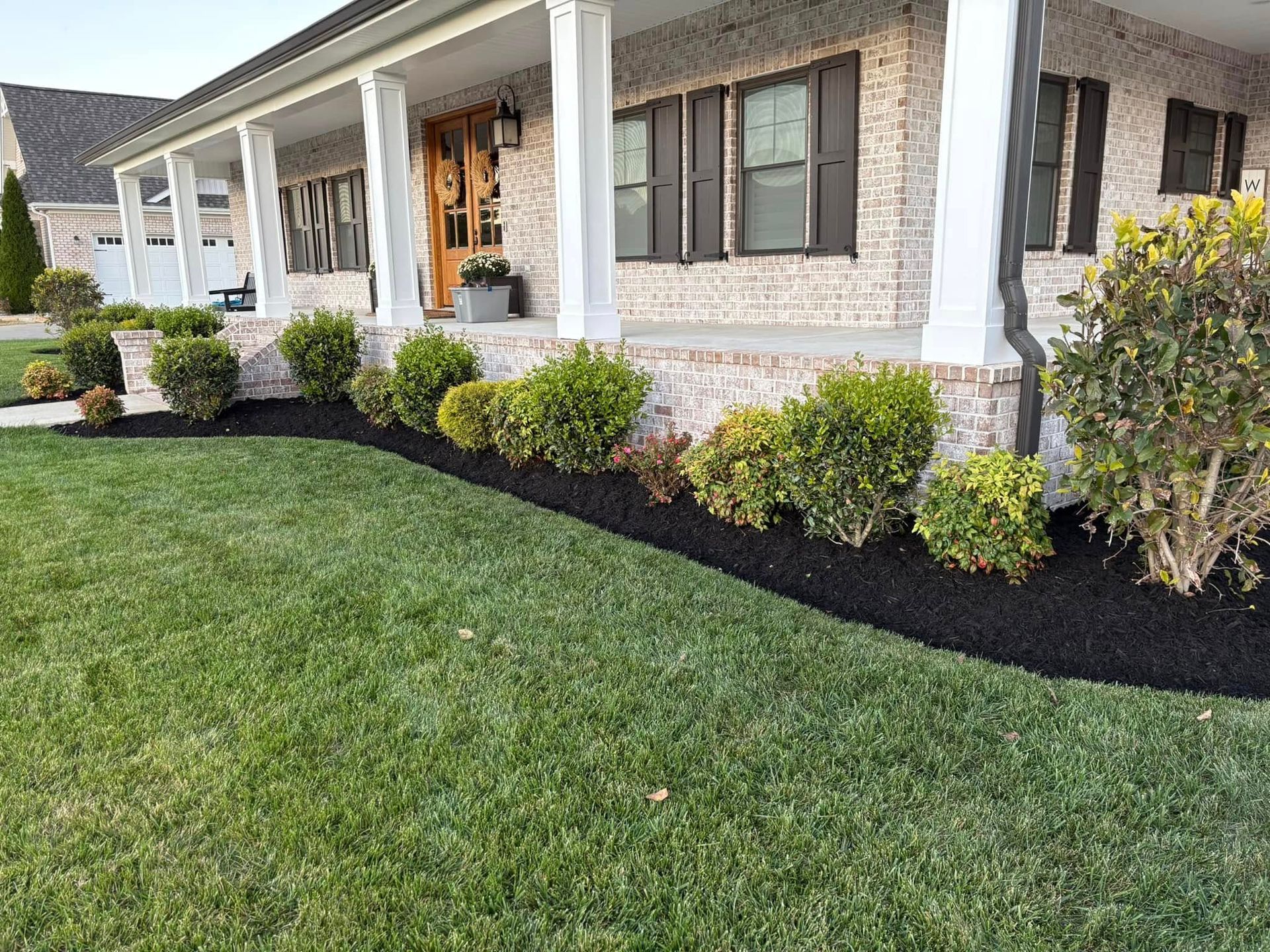 Brick house with white porch, dark shutters, and manicured landscaping with green lawn and black mulch.