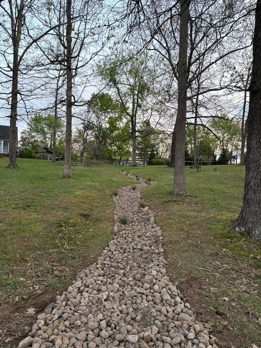 A dry creek bed made of rocks, running downhill through a grassy area with trees.
