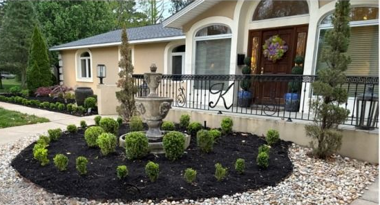 A decorative garden with a fountain, boxwood shrubs, and mulch in front of a house.