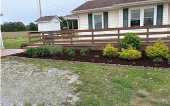 A small beige house with a brown deck, flower bed with various plants, and green lawn.
