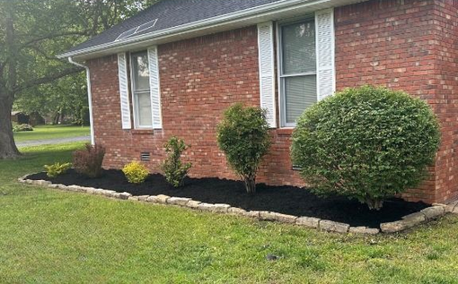 Brick house with a flower bed of plants and black mulch along the front, bordered by stones.