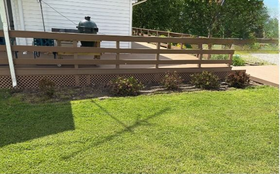 A brown wooden deck with a grill and railing, overlooking a green lawn with some shrubs.