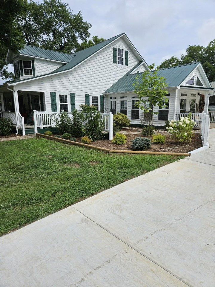 White house with green roof and shutters; front yard with a flower bed and a concrete driveway.
