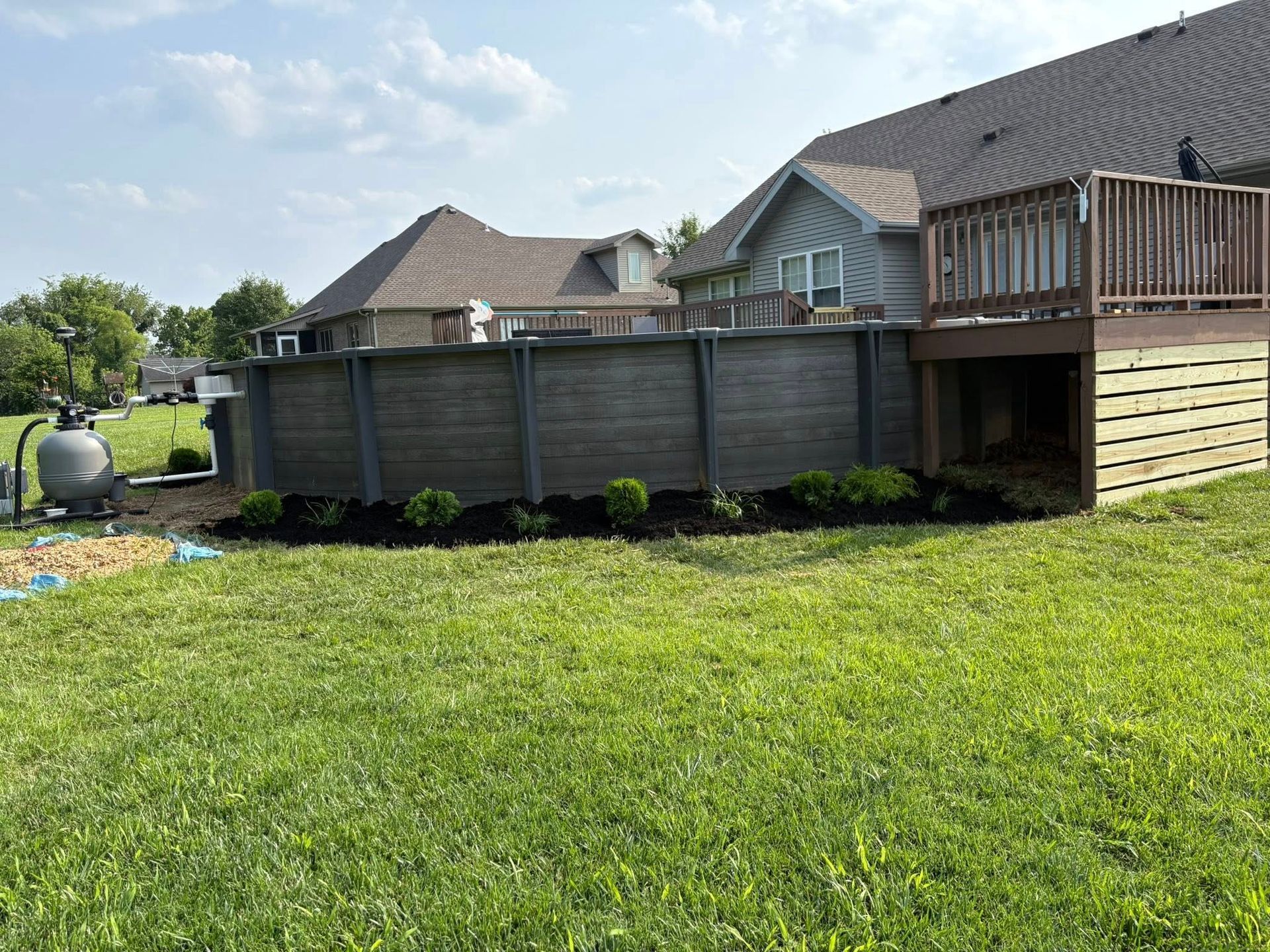 Backyard with above-ground pool and deck, dark gray siding, green lawn, blue sky.