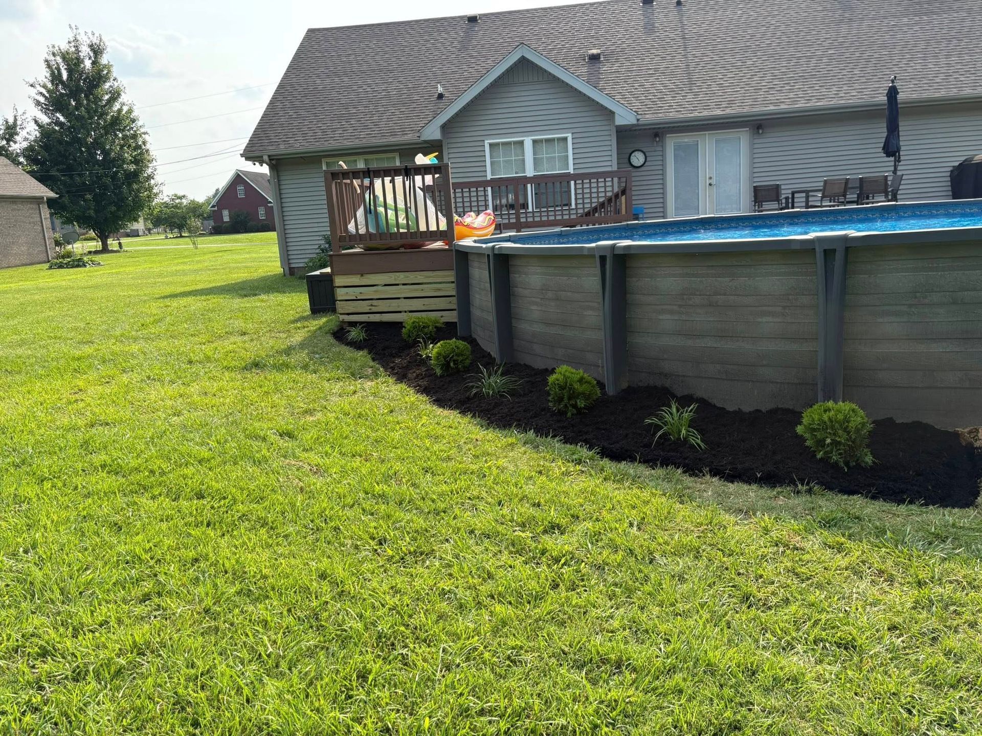 Backyard with an above-ground pool, deck, and landscaped border. Green grass, blue water, and gray siding.