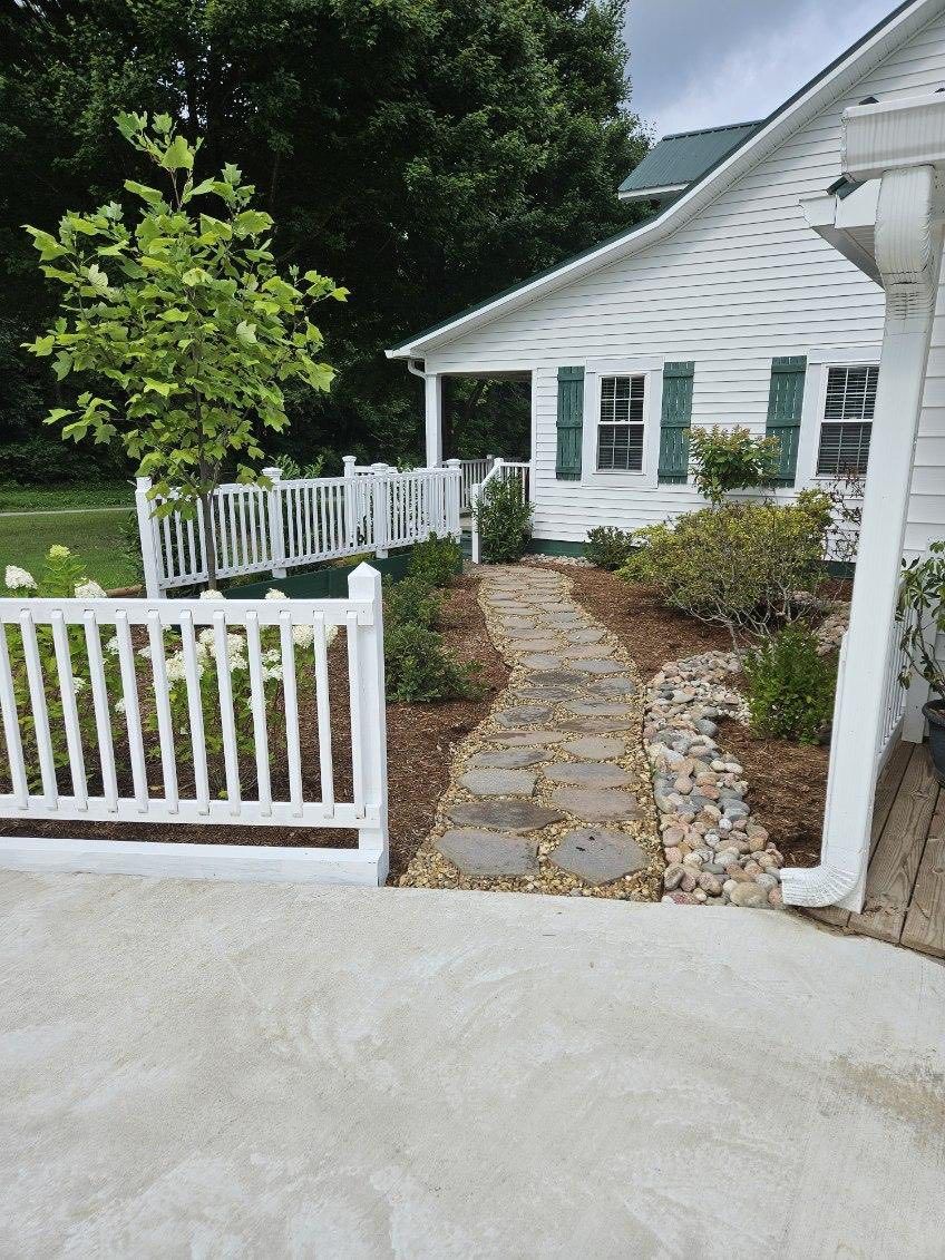 Stone path leading to a white house with green shutters, bordered by white fences and landscaping.