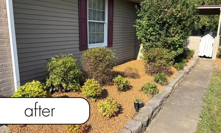 White fence with dark mulch bed, small bushes, brick wall, and blue sky.