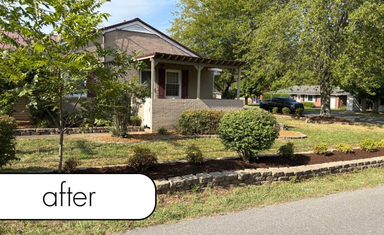White fence with dark mulch bed, small bushes, brick wall, and blue sky.