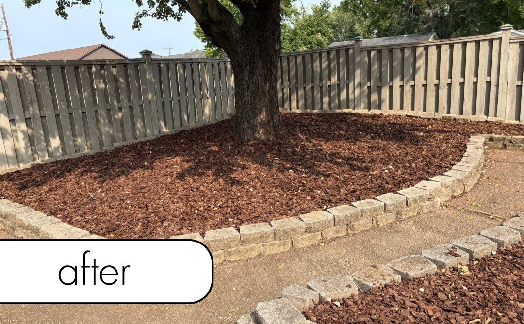 White fence with dark mulch bed, small bushes, brick wall, and blue sky.