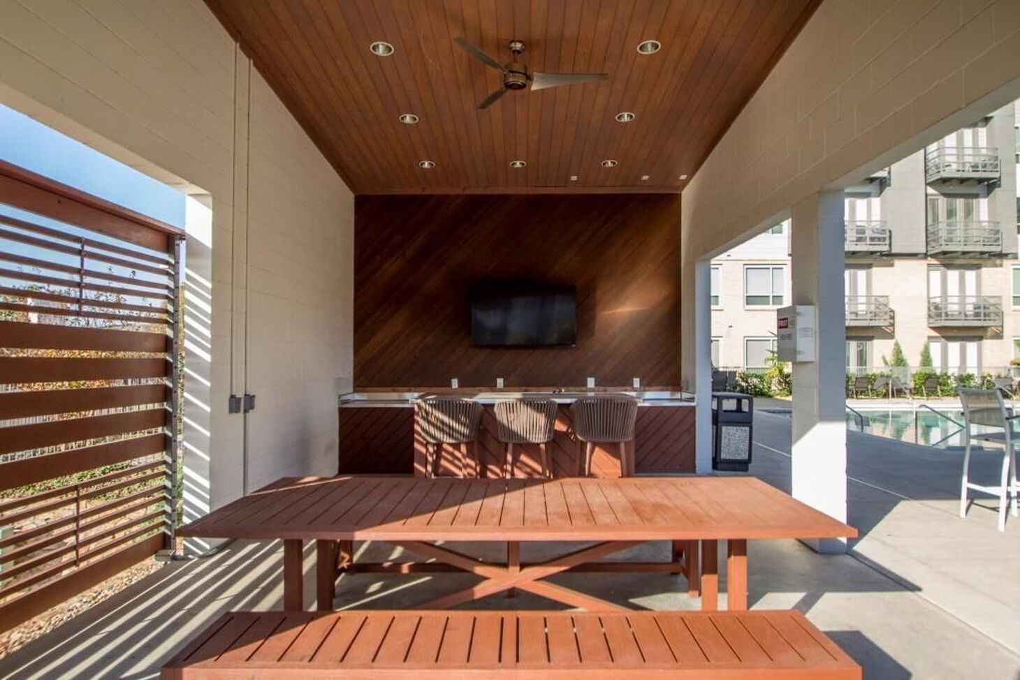 Outdoor grilling area with wooden table, benches, and built-in grill, next to a pool.