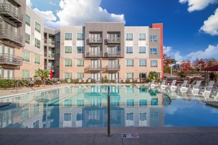 Apartment building with pool on a sunny day. Building is gray with balconies. Pool has reflections.