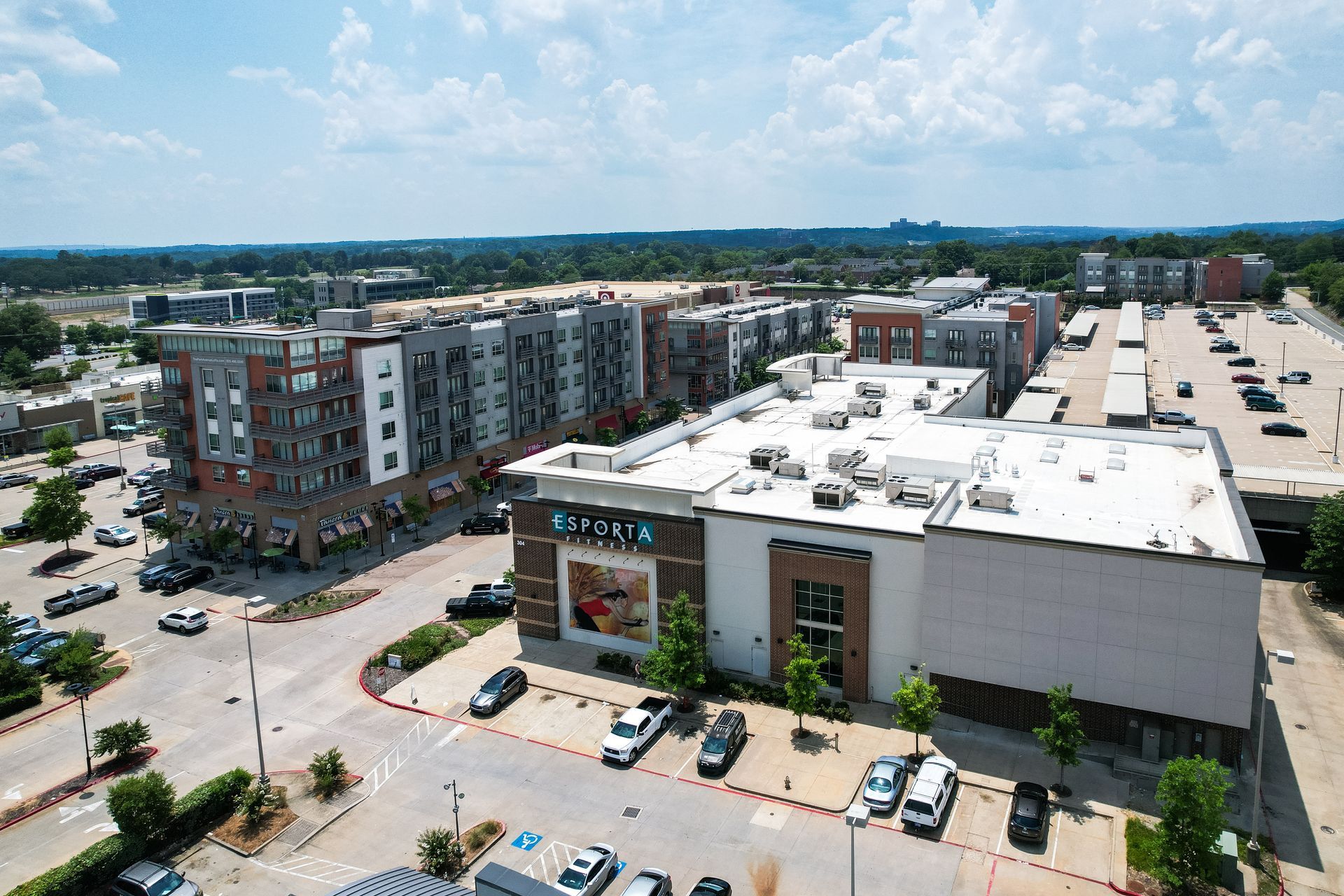 Exterior aerial view of a shopping center with multi-story apartments and a parking lot on a sunny day.