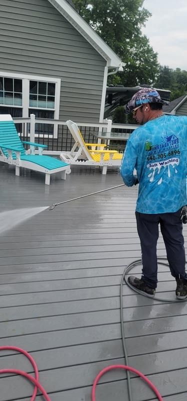 A person power washes a gray deck, with lounge chairs nearby. They wear a hat and blue shirt.