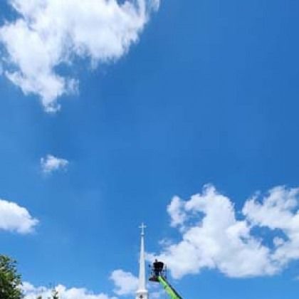 Church steeple against a bright blue sky with puffy white clouds; a person on a lift appears to be working on the steeple.