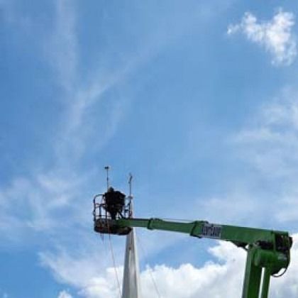 Person in a lift basket repairing something on a tall pole against a blue sky with clouds.