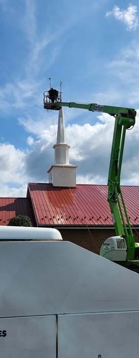 A lift platform with a person working on a church steeple under a blue cloudy sky.