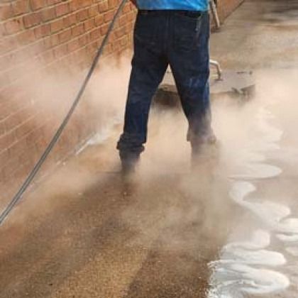 Person power washing a concrete surface, creating a cloud of mist, near a brick wall.