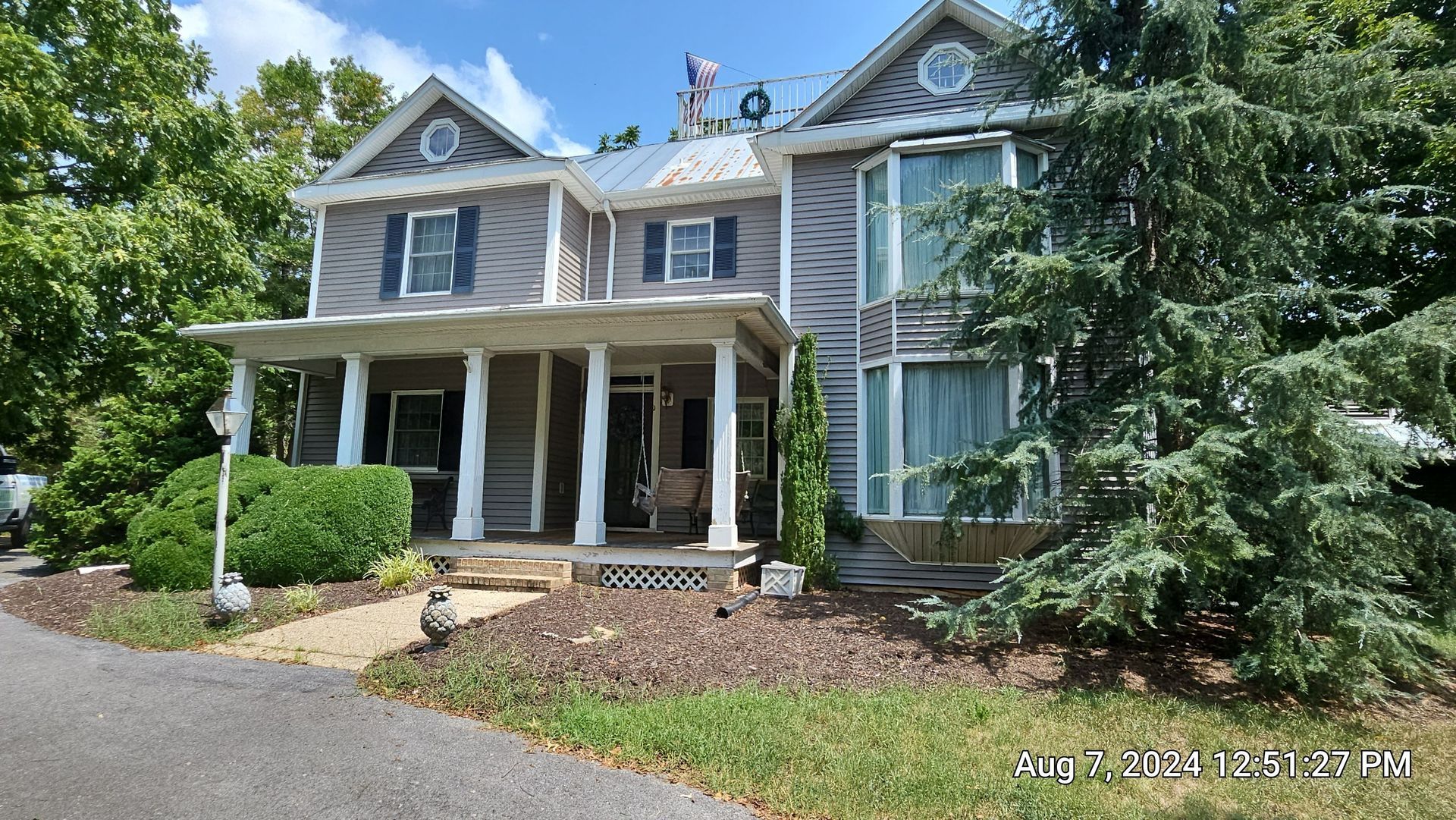 Grey, two-story house with porch and columns, surrounded by trees and a paved driveway.