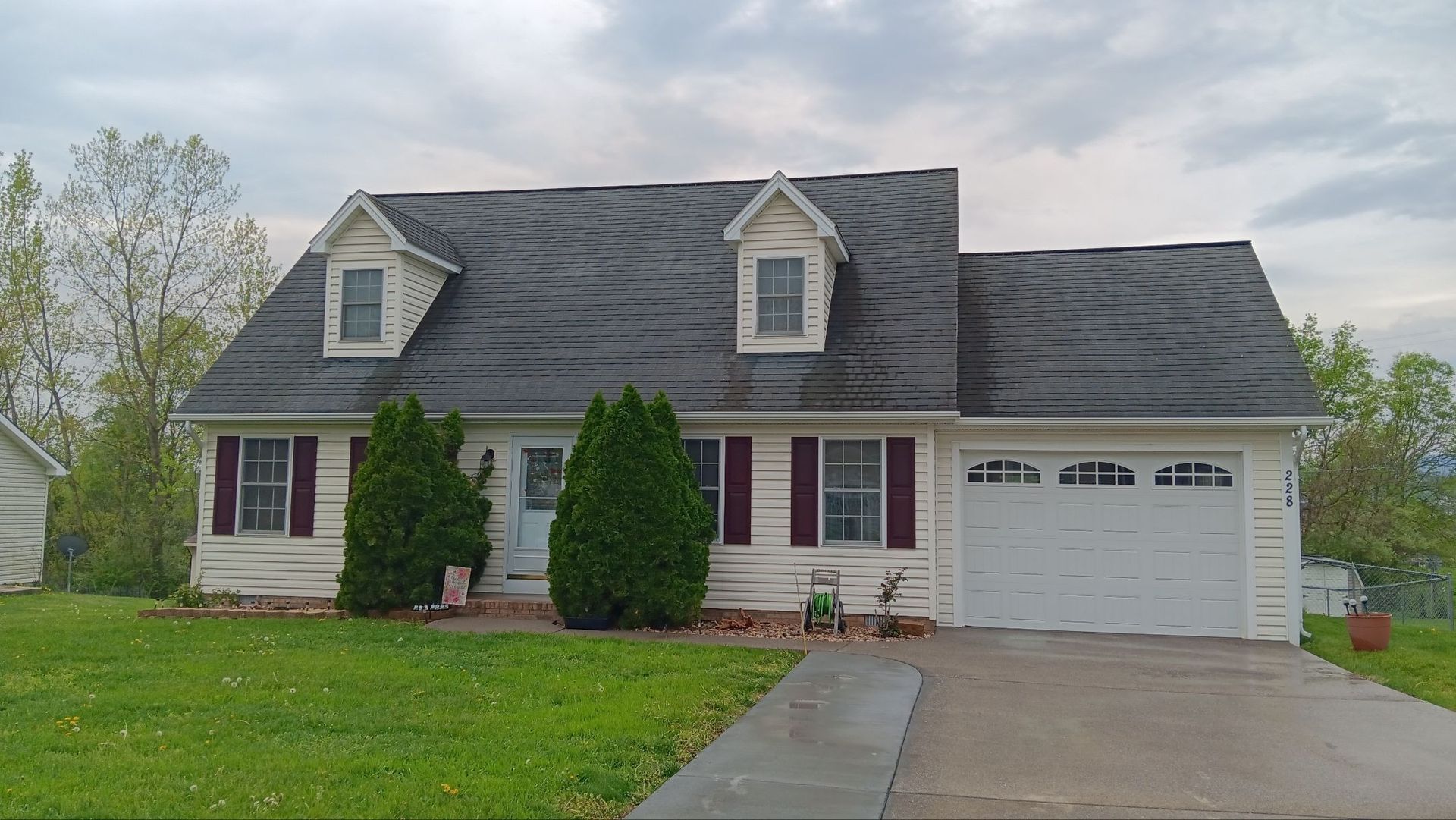 White house with dark roof and red shutters, green lawn and driveway, cloudy sky.