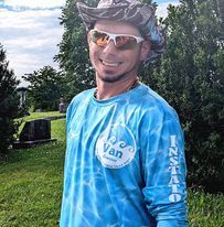 Man wearing sunglasses, hat, and blue tie-dye shirt smiles outdoors near a grassy area.