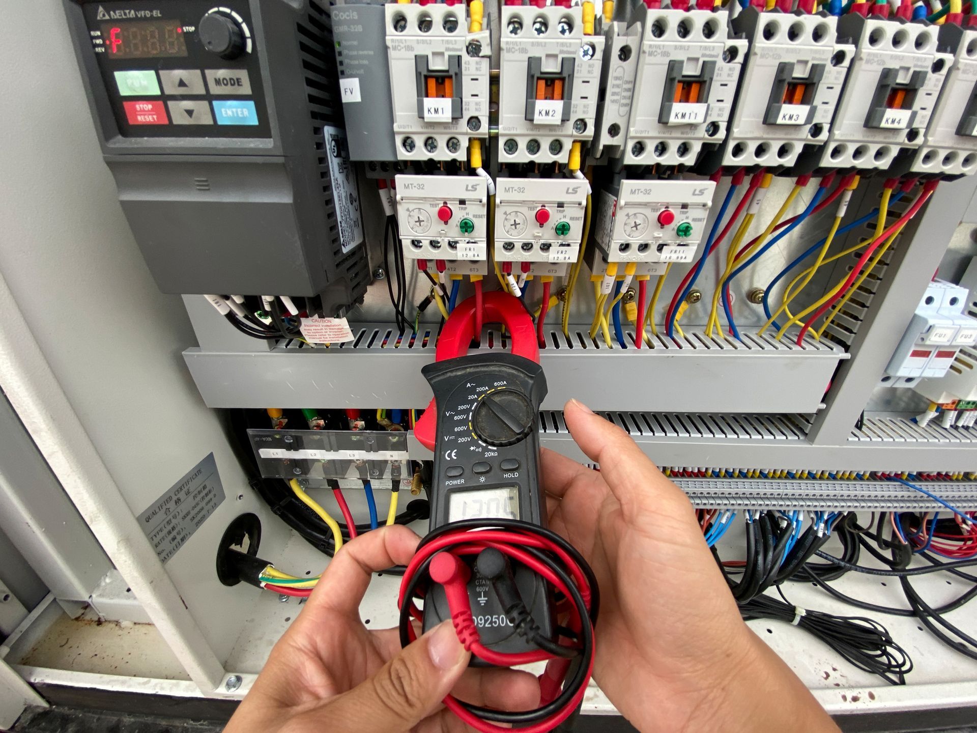 Hands holding a multimeter to measure electrical current in an industrial control panel with wires and components.