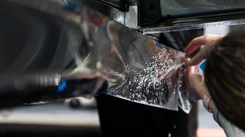 Person applying a clear protective film to a car's surface, indoors.