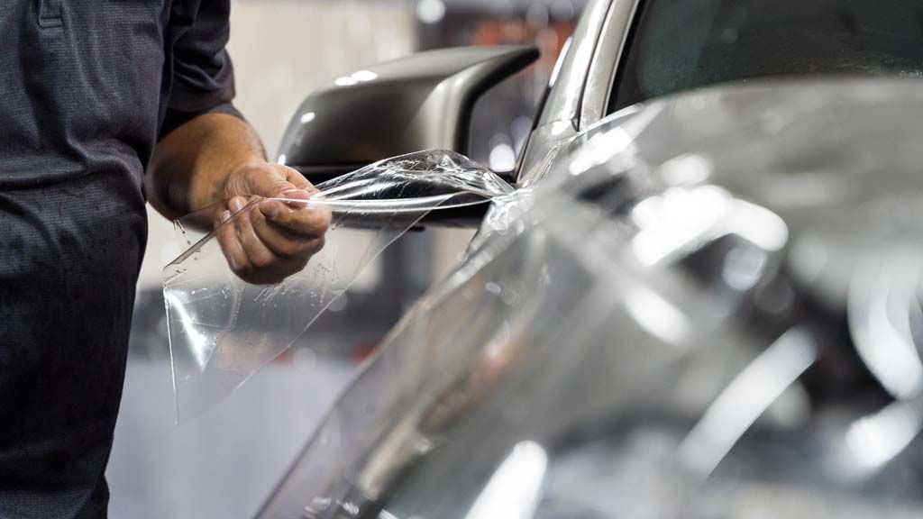 Person applying protective film to a car's side panel; the car is silver.