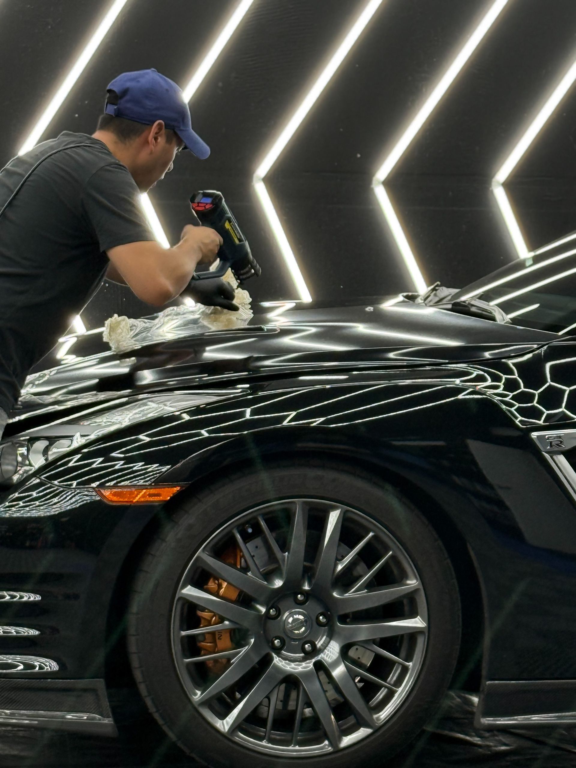 Man waxing black car's hood in well-lit garage. The car's orange headlight is visible.