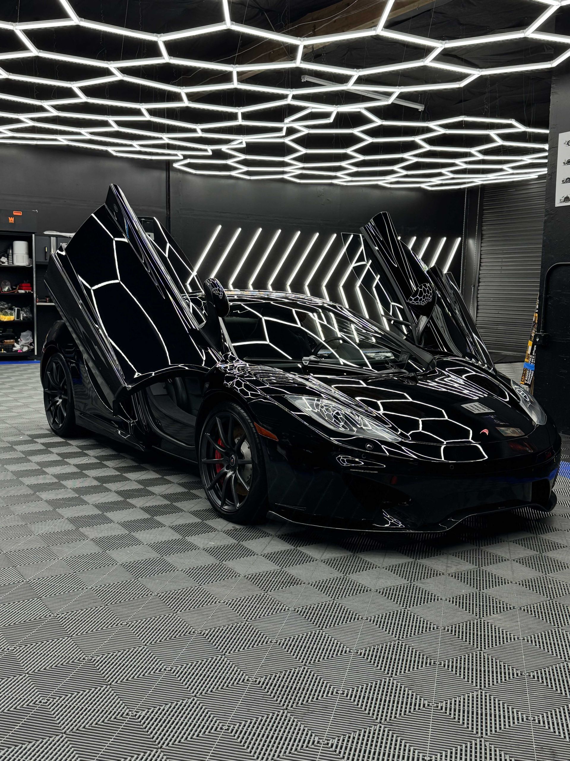 Black Lamborghini with doors up, in a garage with hexagon ceiling lights and checkered floor.